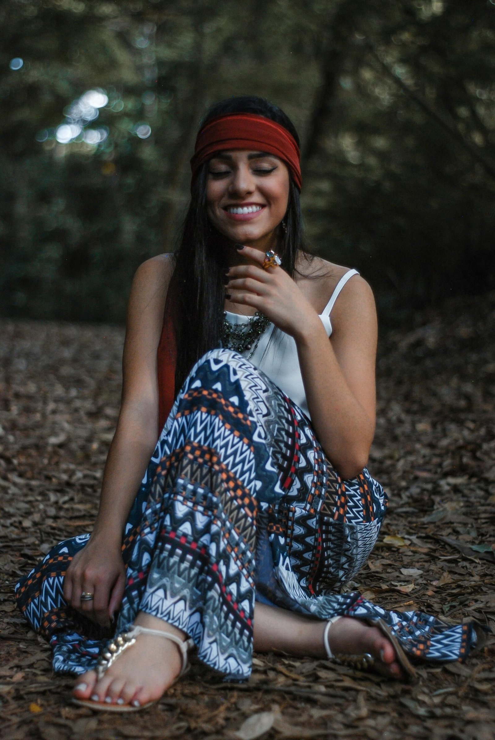 A young woman with a bohemian style outfit enjoys a peaceful moment in nature, sitting on leaf-covered ground.