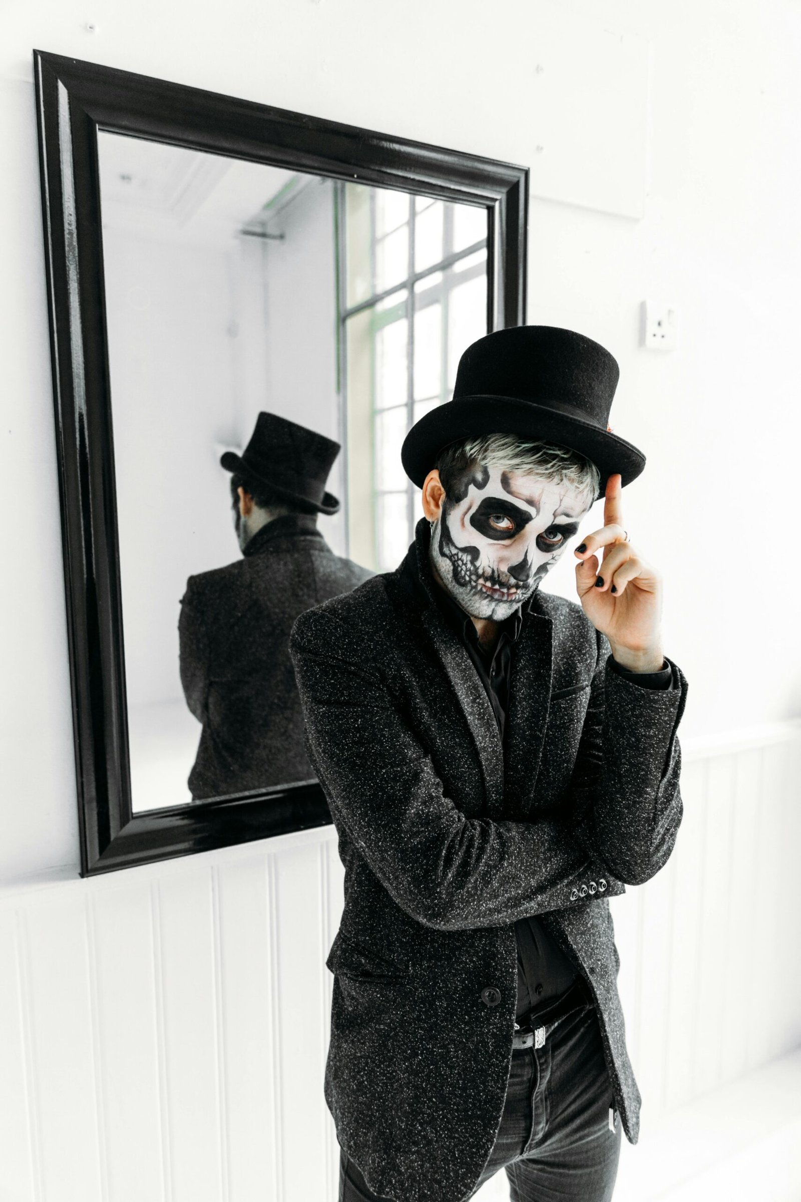 Man in artistic skull makeup with top hat standing in front of a mirror, exuding a mysterious vibe.